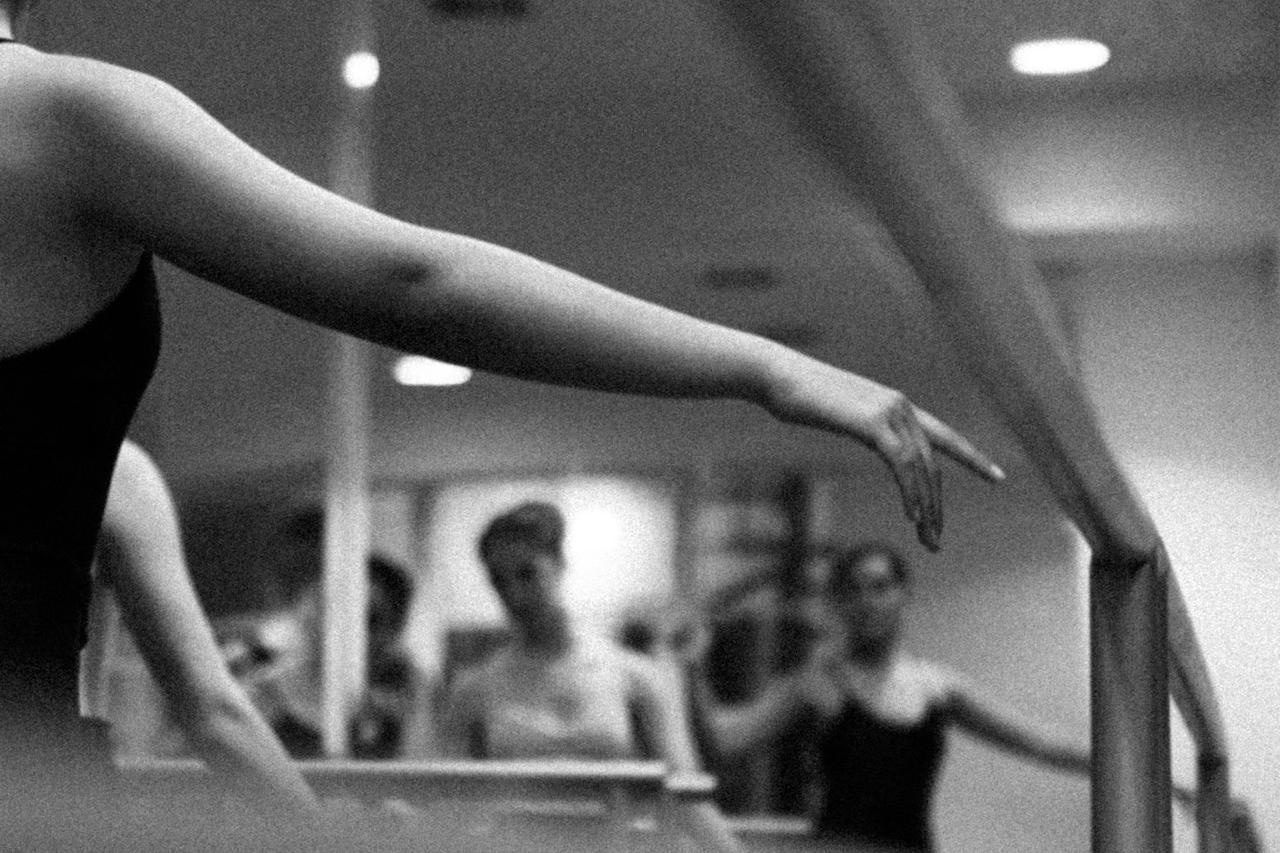 El brazo y la mano de una bailarina sobre la barra, con la clase desenfocada al fondo