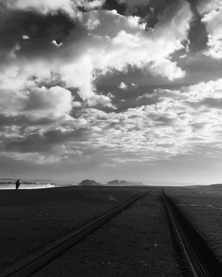 Playa con huellas de neumáticos, un pescador y un cielo dramático de nubes