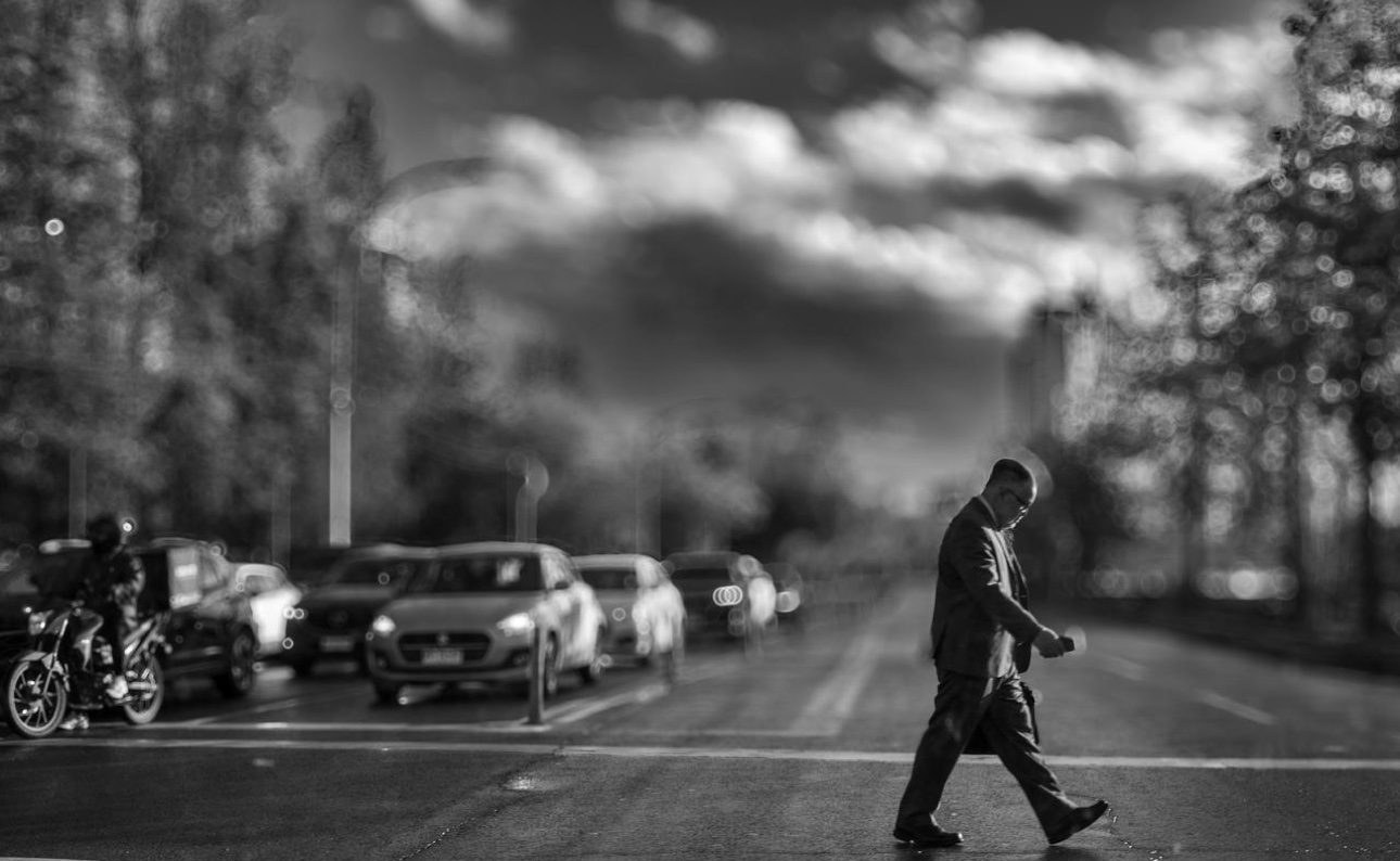Hombre de traje cruzando la calle mirando su teléfono, bajo un cielo dramático de nubes
