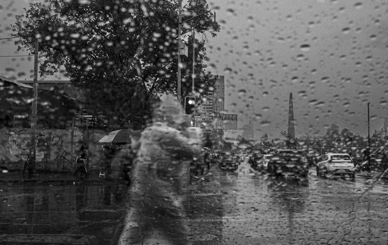 Una avenida bajo la lluvia, vista a través del parabrisas con gotas, con una figura borrosa cruzando