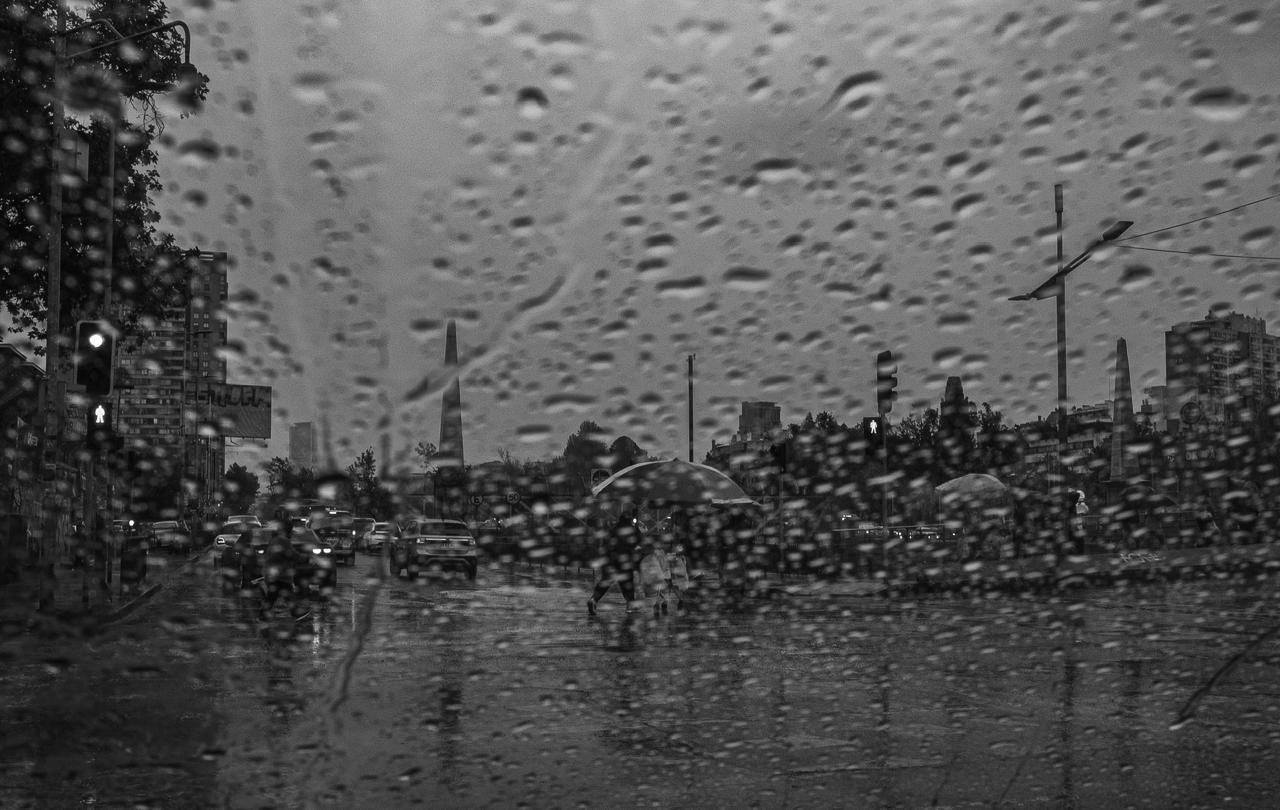 Una persona con paraguas cruzando la avenida bajo la lluvia, con los obeliscos al fondo, todo a través del vidrio con gotas